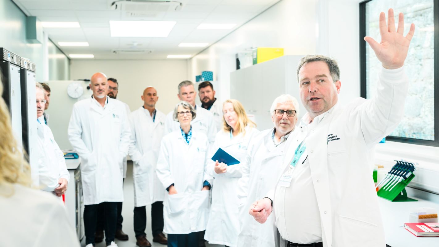11 senior scientists in lab coats examining a new laboratory with one in foreground gesturing into the distance